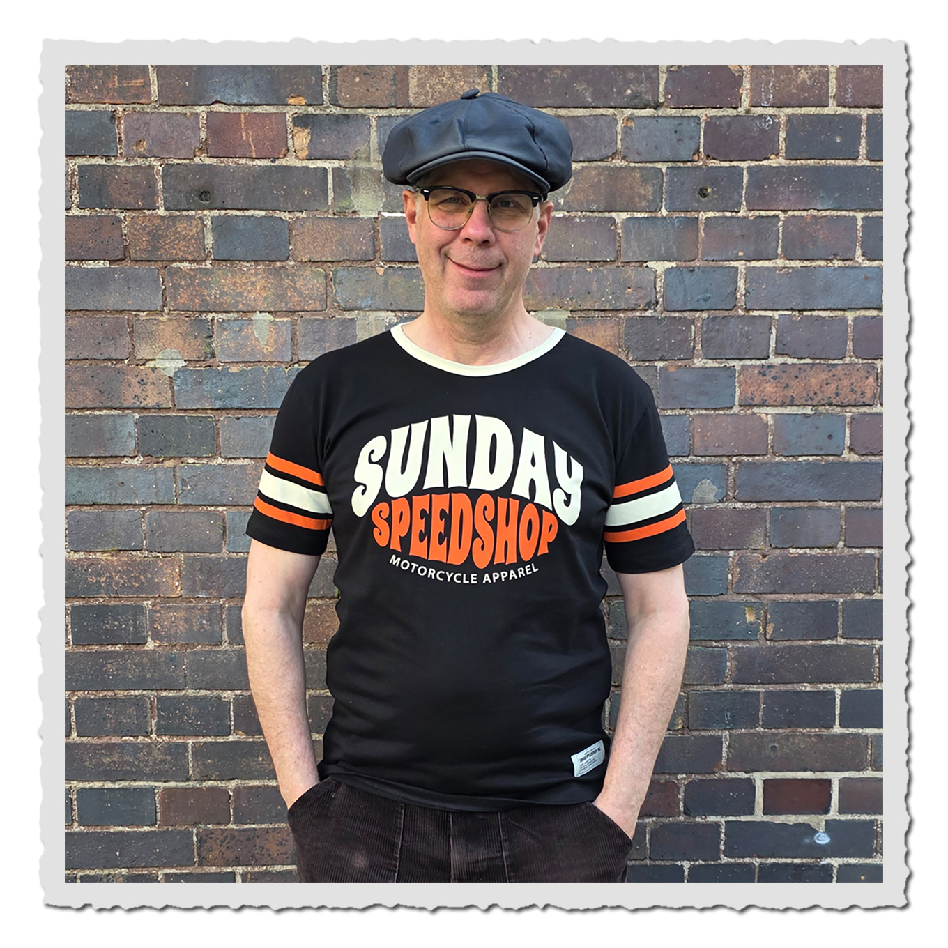 Person von vorn mit schwarzem Sunday Speed Shop Shirt vor gemauerter Backsteinwand.